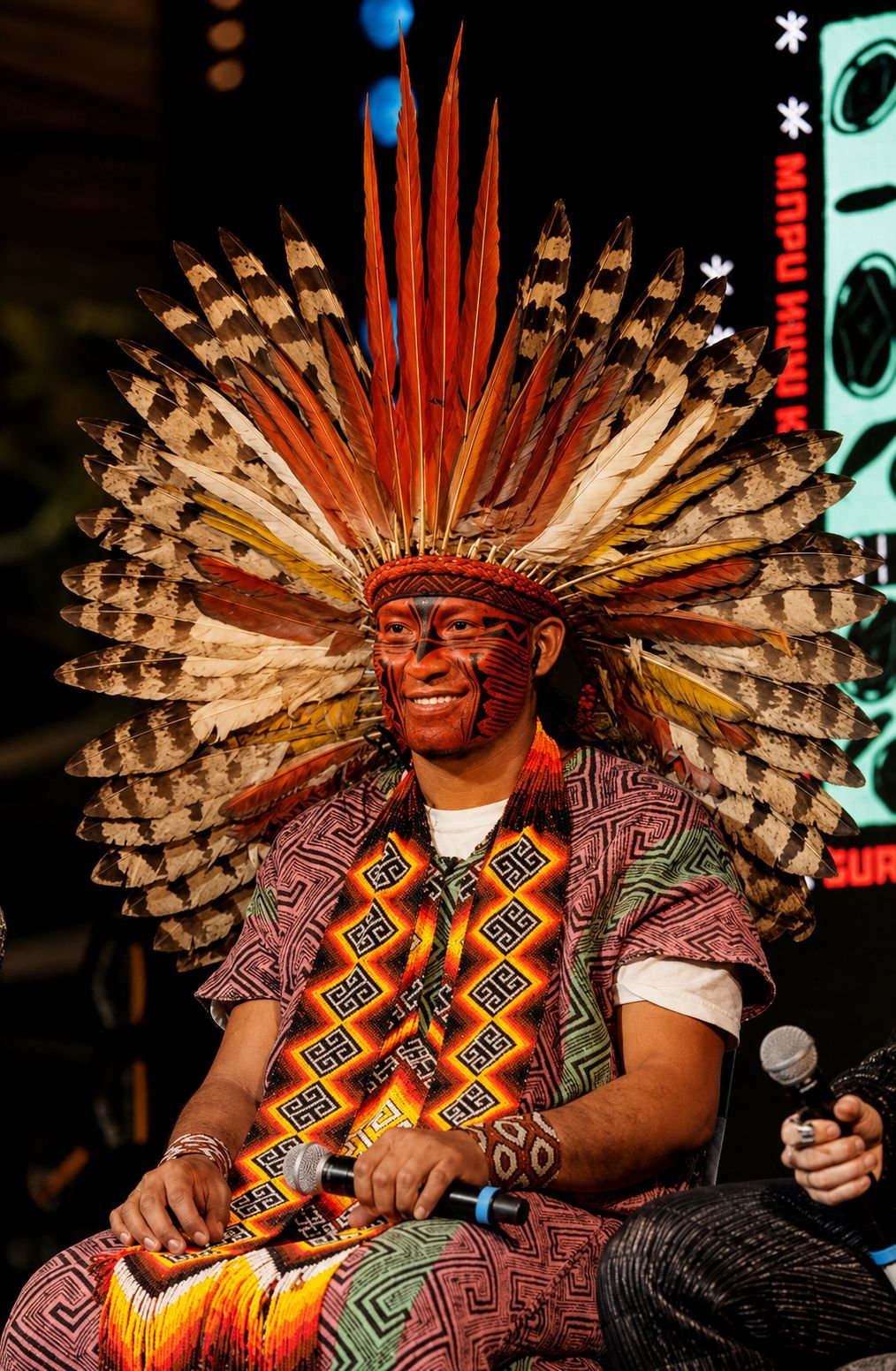Mapu Huni Kuin sentado, com cocar de penas e veste tradicional estampada, durante uma apresentação