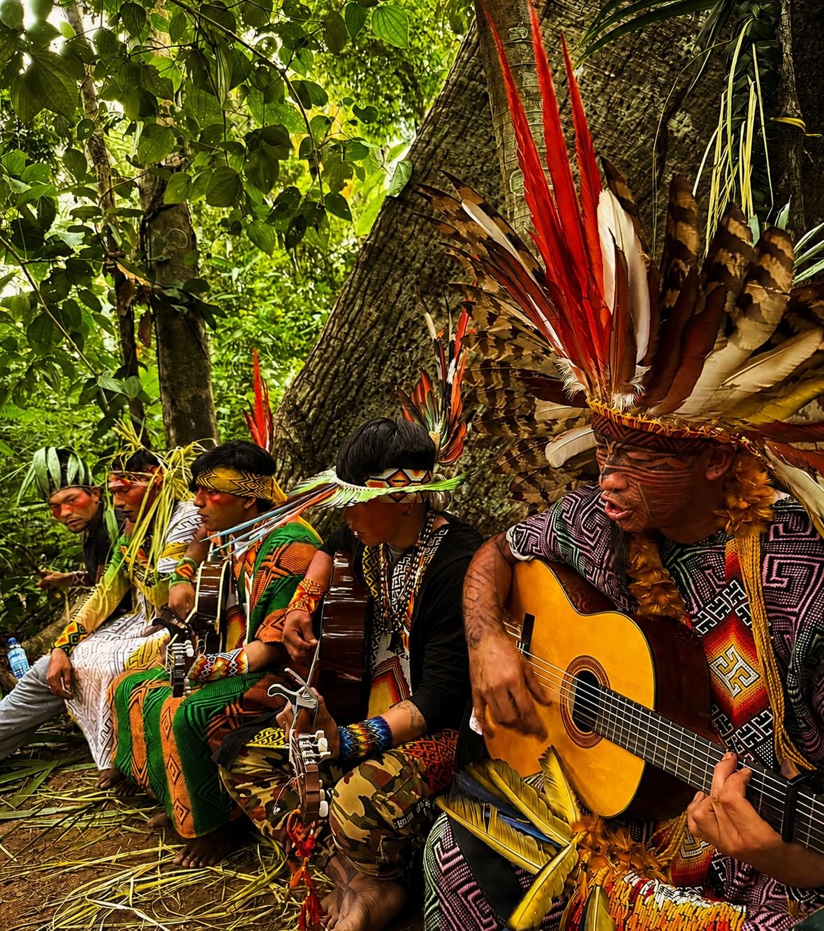 Mapu Huni Kuin toca violão em roda com parentes na floresta, usando cocar de penas